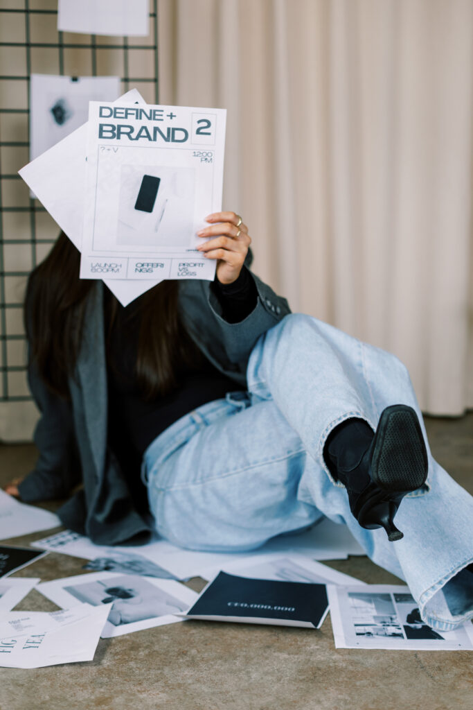 Woman seated among printed brand concepts while holding a page labeled “Define + Brand.”