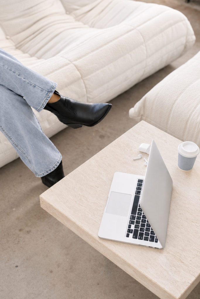 Person in pale blue jeans and black boots seated beside an open laptop, coffee cup, and wireless earbuds.