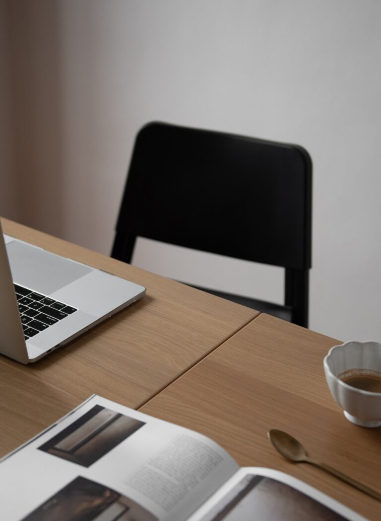 Laptop, coffee, and an open magazine on a wooden table beside a black chair.