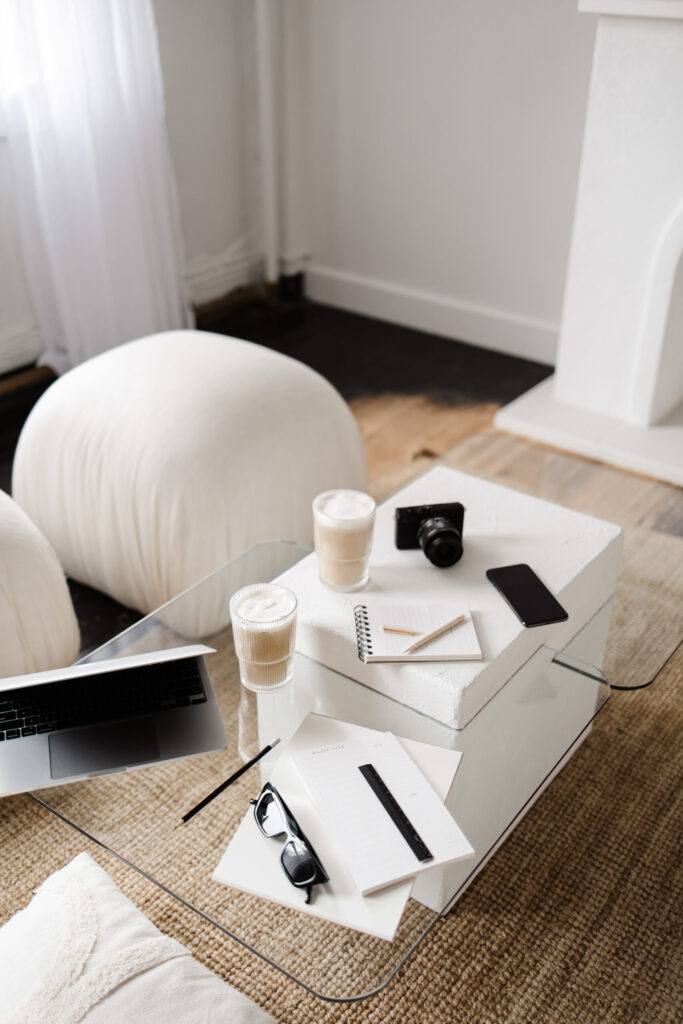 Glass coffee table arranged with a laptop, camera, smartphone, notebooks, pencils, glasses, and two drinks.