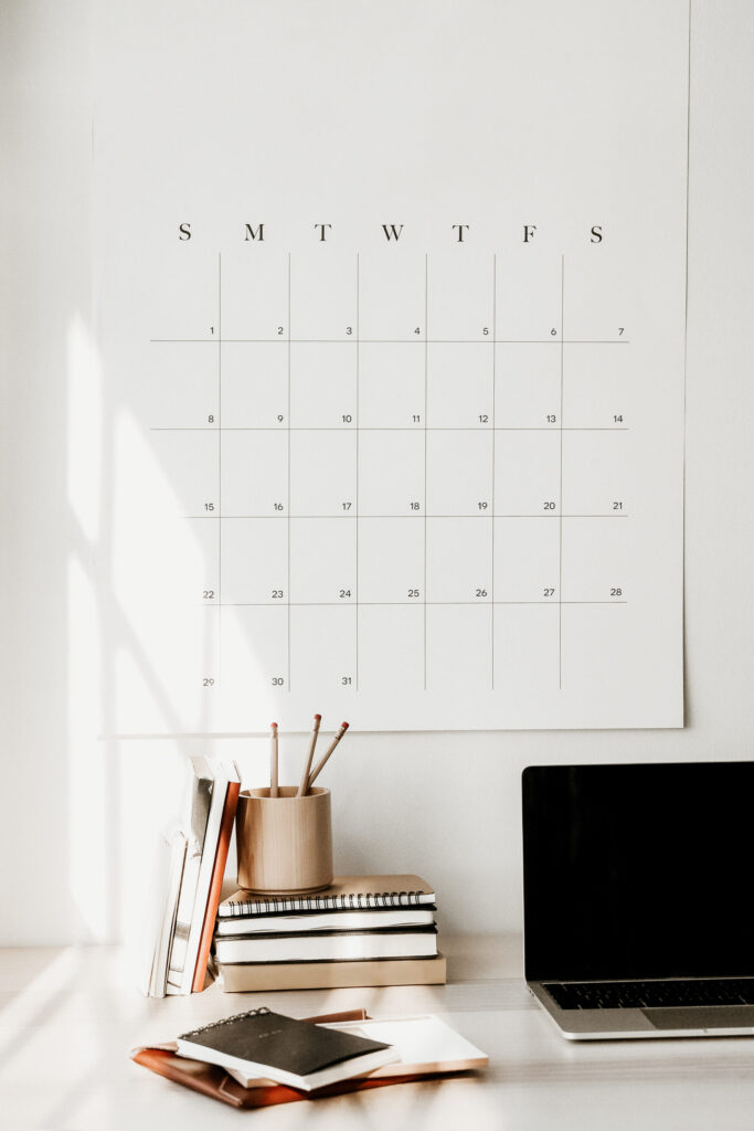 A large monthly wall calendar above a desk with an open laptop, stacked notebooks, books, and a wooden pencil cup.