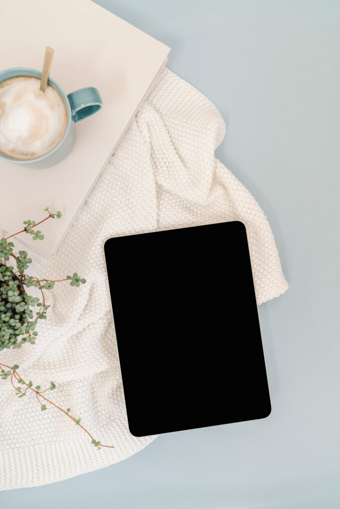 A tablet with a blank black screen beside a blue coffee mug, cream knitted blanket, closed laptop, and trailing plant.