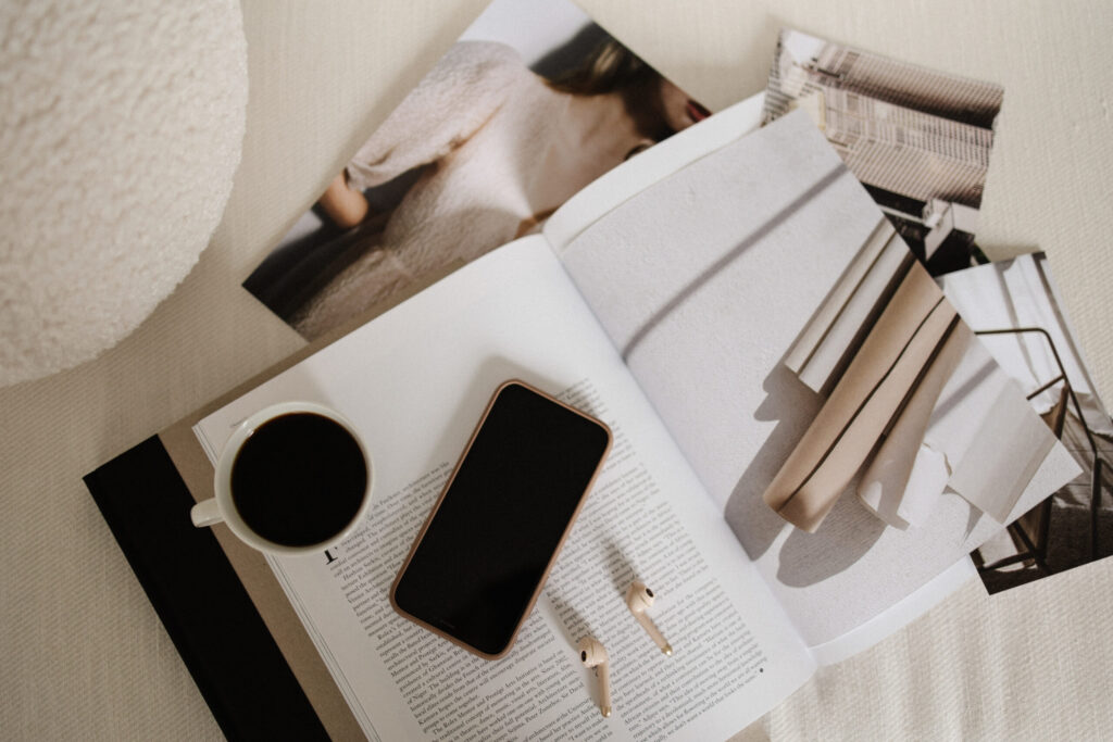 Coffee, smartphone, wireless earbuds, an open magazine, and printed photographs arranged on cream fabric.