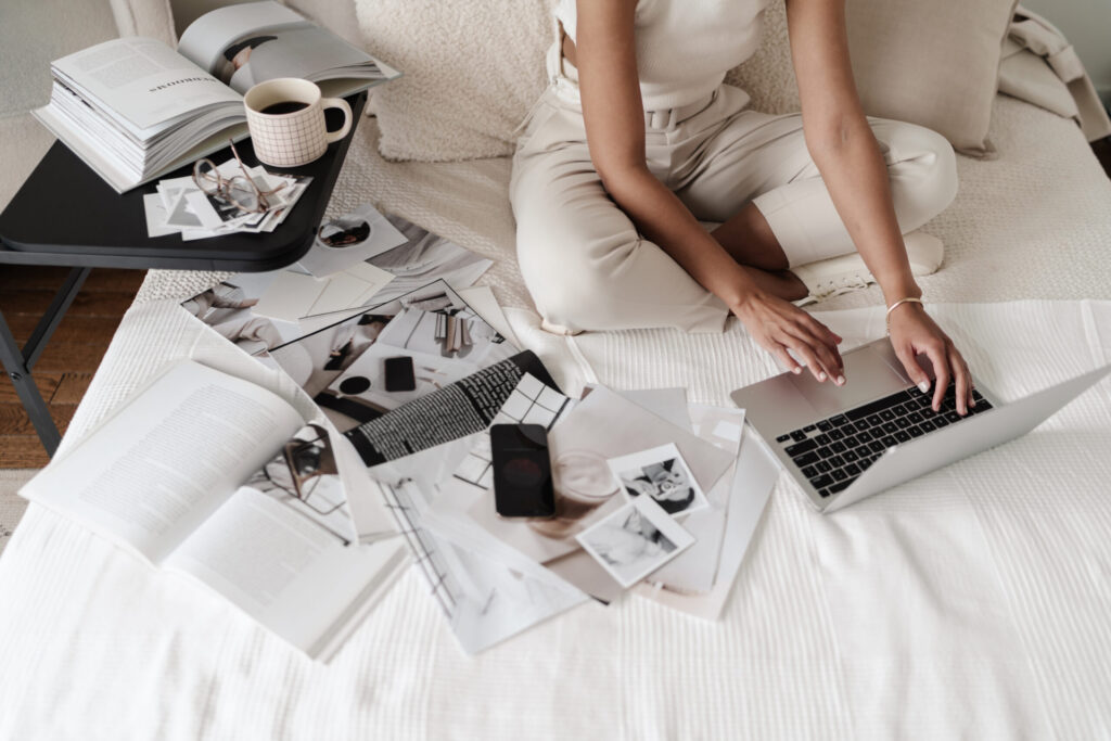 Woman using a laptop on a cream bed surrounded by open magazines, printed photographs, a smartphone, and coffee.