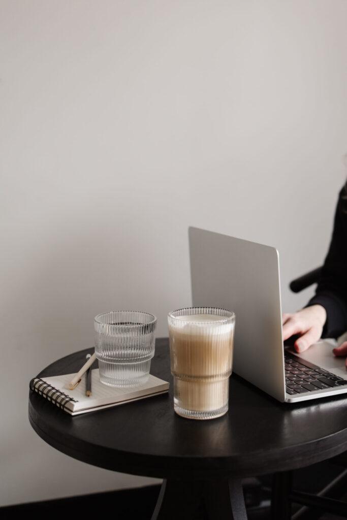 Person working on a laptop at a round black table with a notebook, pens, water, and an iced coffee.