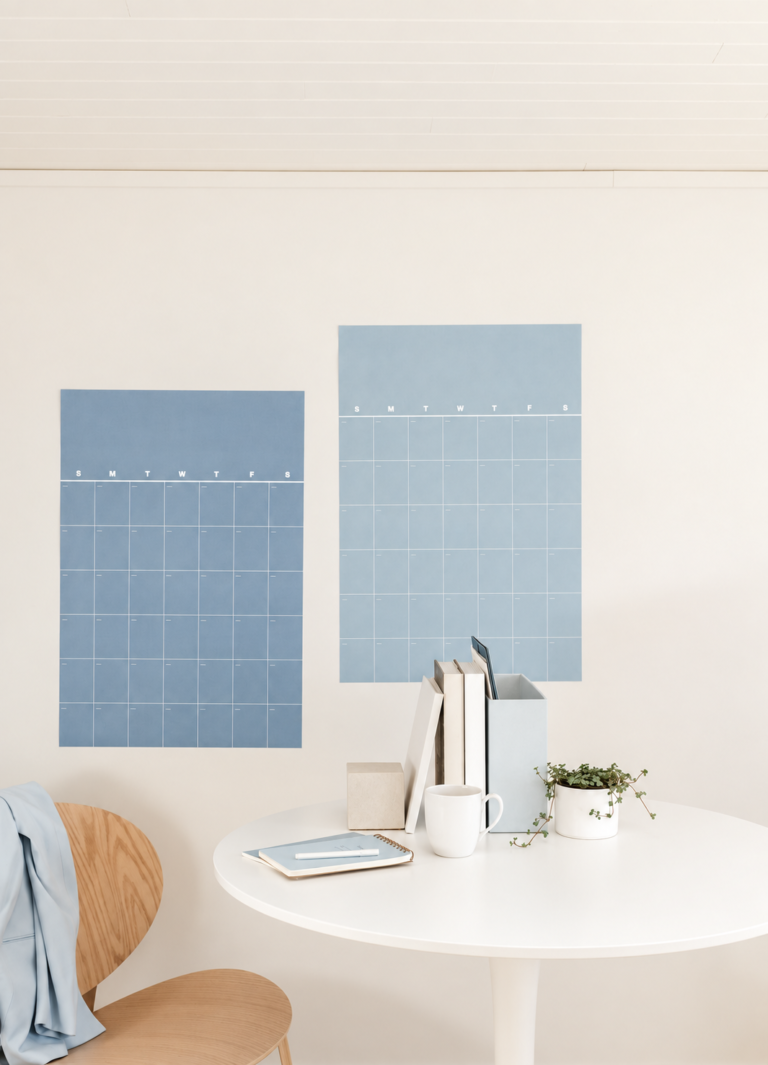 Two powder-blue wall calendars above a round white desk with books, a notebook, a mug, and a trailing plant.