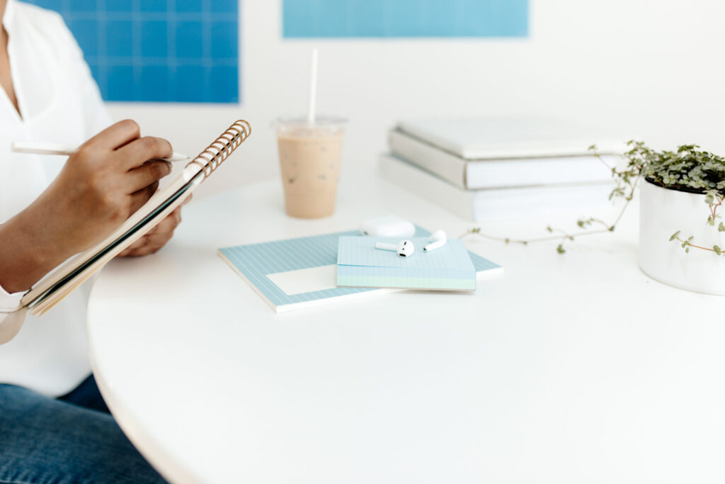A person writing in a spiral notebook at a white desk with blue stationery, iced coffee, books, and a small plant.
