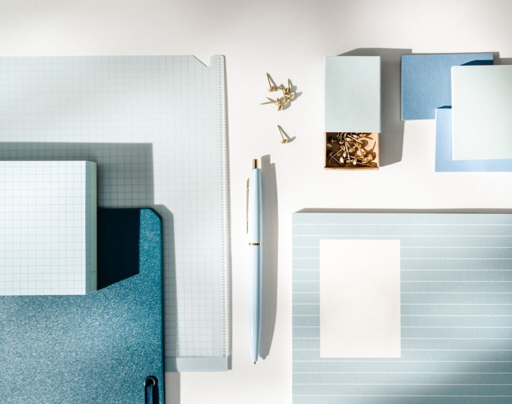 Blue file folders, grid paper, note cards, a pen, and gold thumbtacks arranged on a white surface.