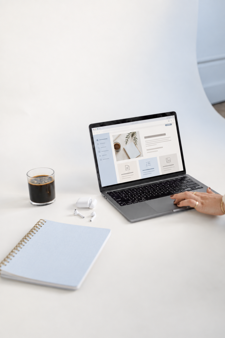 Laptop displaying a creative business website beside a notebook, coffee, and wireless earbuds on a pale studio desk.