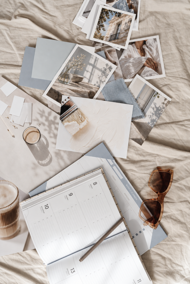 Open planner, tortoiseshell sunglasses, coffee, powder-blue paper samples, fabric, and editorial photographs arranged on cream linen.