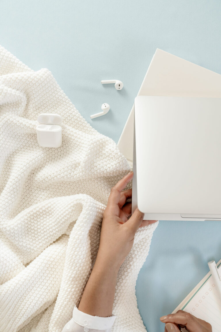 An open silver laptop on a powder-blue surface beside a white knitted blanket, wireless earbuds, and a checklist notebook.