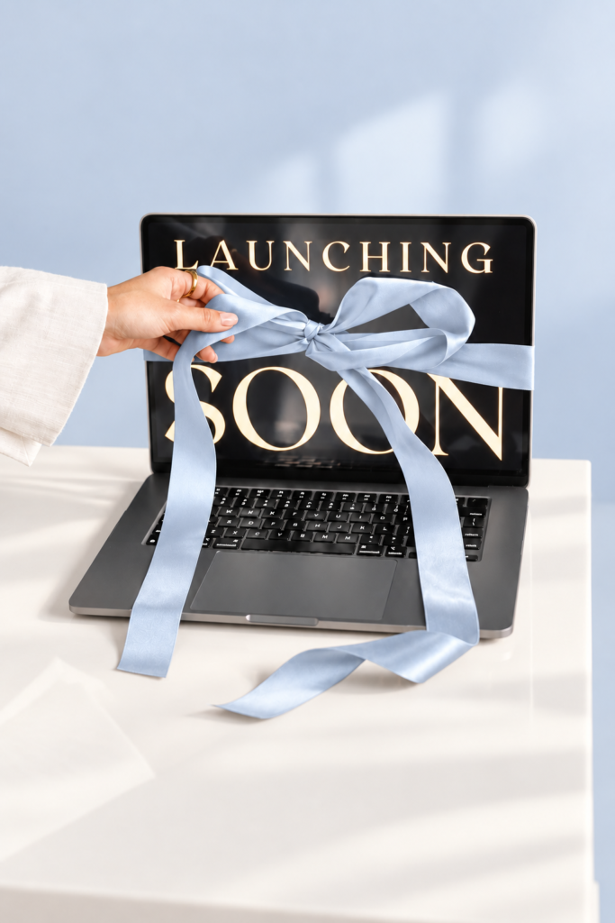 Hand untying a navy satin ribbon wrapped around a laptop displaying “Launching Soon.”