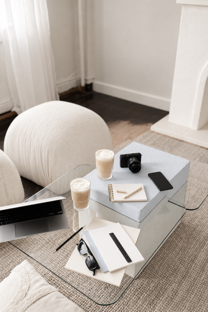 Creative workspace with a laptop, camera, phone, notebooks, coffee, and glasses arranged on a glass table.