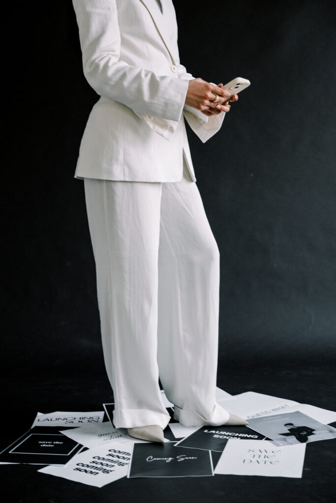 Business owner in a white suit checking her phone while standing among printed launch announcements.