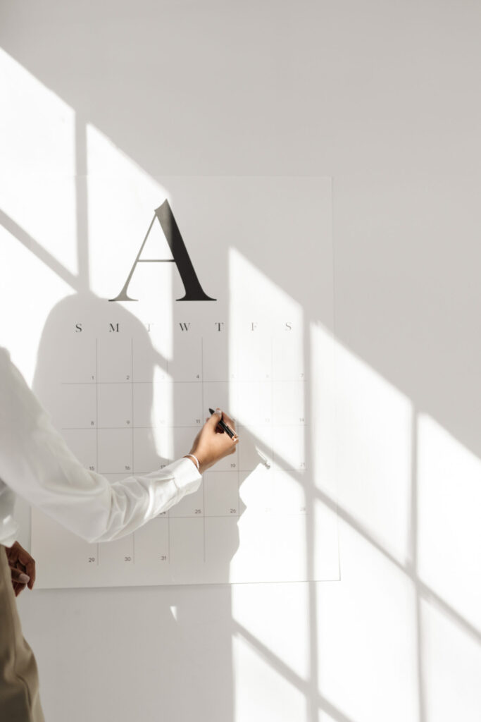 A person writing on a large monthly wall calendar in a sunlit office.