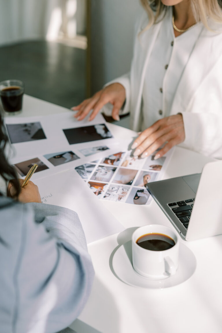 Two women review a printed photo layout beside a laptop and cups of coffee.