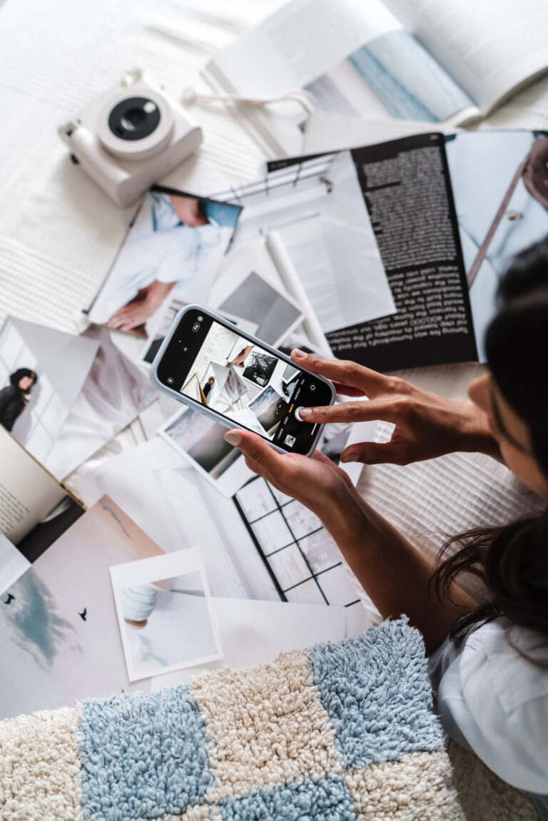 A business owner photographs printed brand images with a phone beside an open laptop and instant camera.