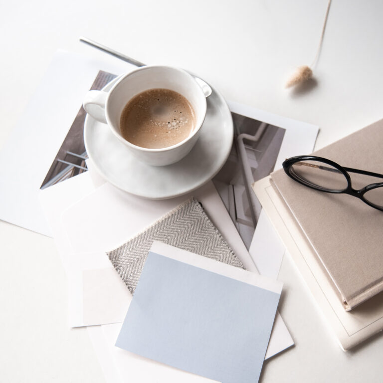 Coffee, color cards, printed photographs, a fabric swatch, glasses, and a notebook arranged on a white desk.