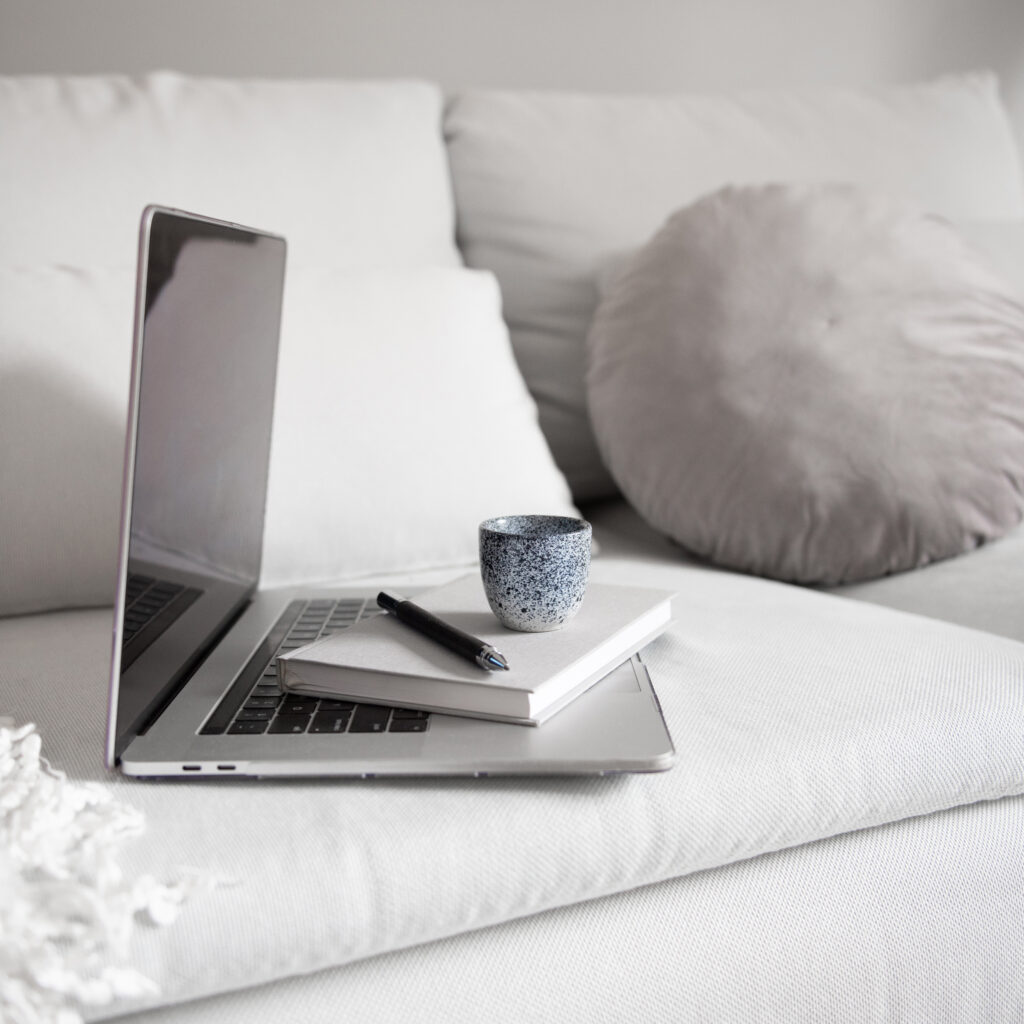 An open laptop with a notebook, pen, and ceramic cup arranged on a neutral sofa.