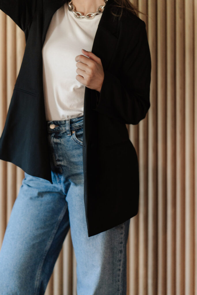 A woman wearing a black blazer over a white top with light-wash jeans and a silver chain necklace.