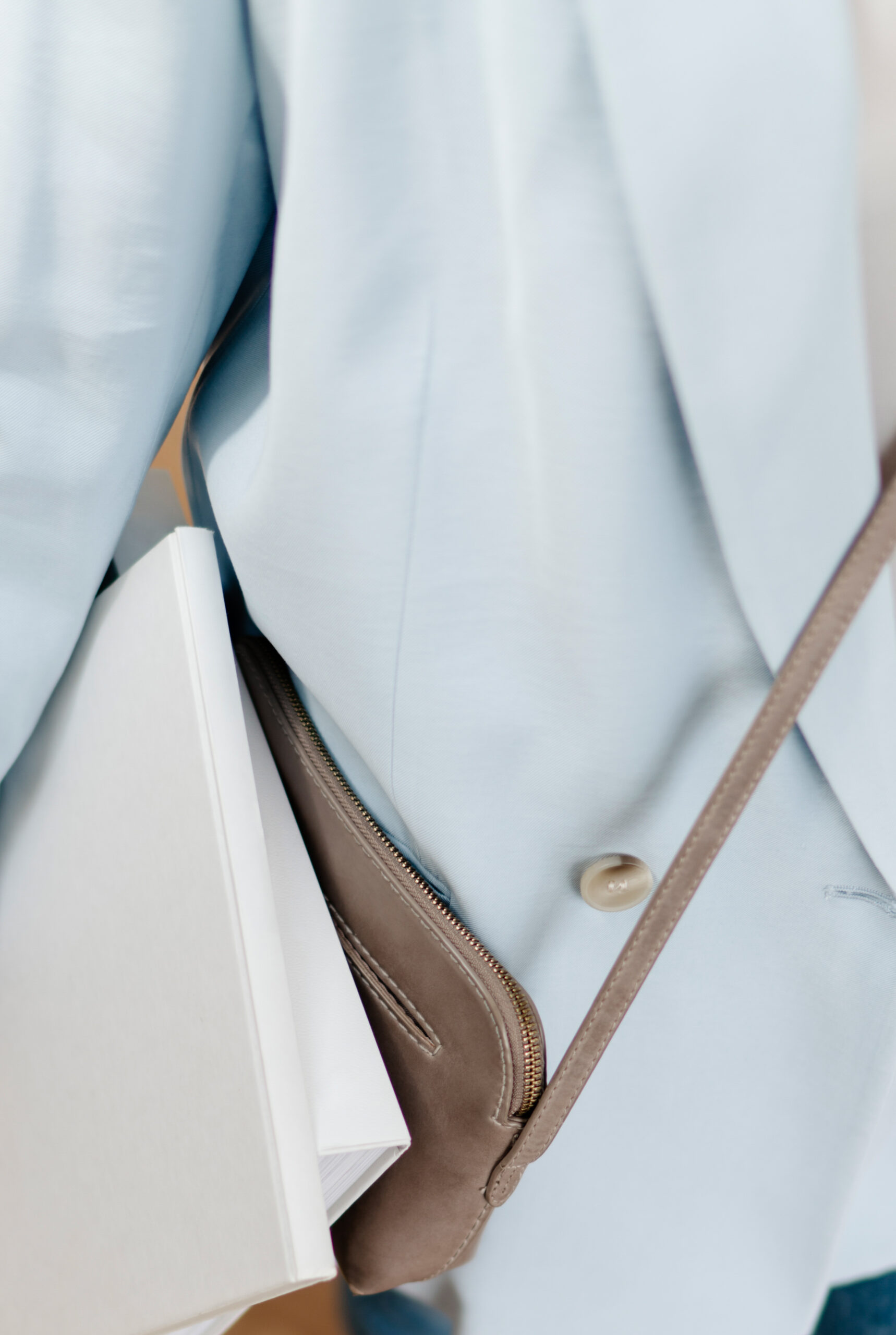 Close-up of a pale blue blazer styled with a taupe crossbody bag and white notebook.