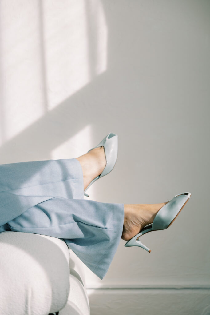 A person wearing pale blue trousers and matching open-toe heels while seated in a sunlit room.
