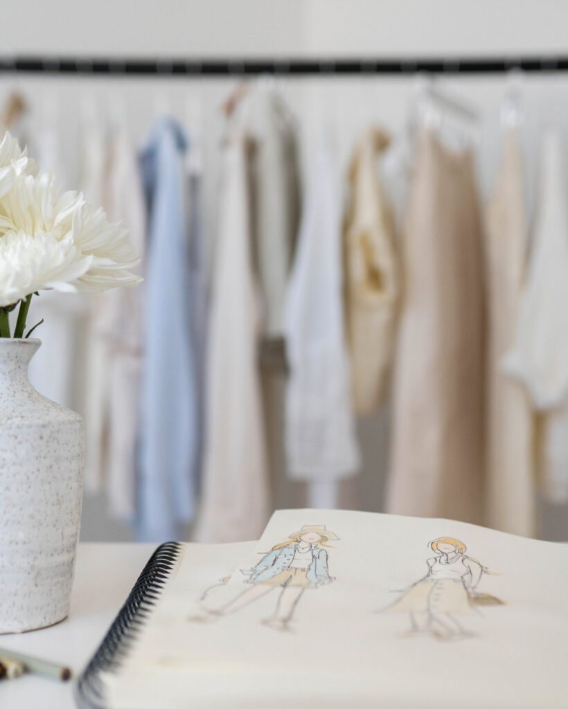 Fashion sketches and white flowers on a desk with neutral and blue clothing hanging in the background.