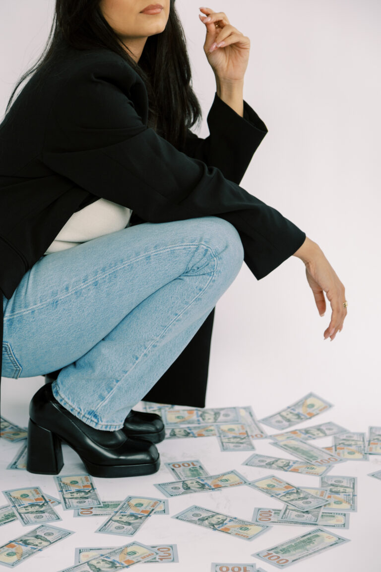 A woman in a black blazer and jeans poses among scattered dollar bills against a light background.