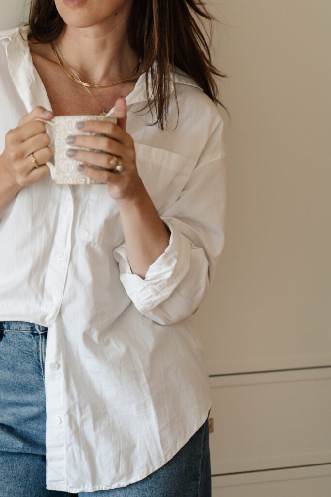 A woman in a white shirt and jeans holding a textured ceramic mug against a warm neutral wall.
