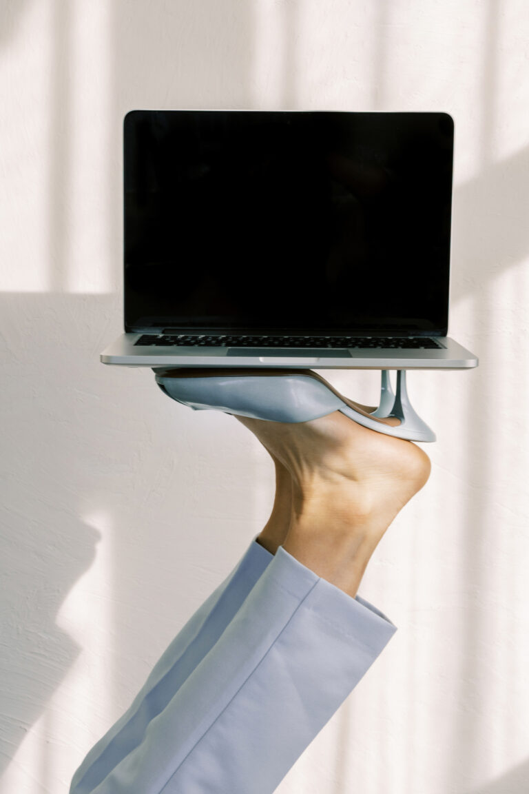 An open laptop balanced on a pair of blue high heels against a softly lit neutral background.