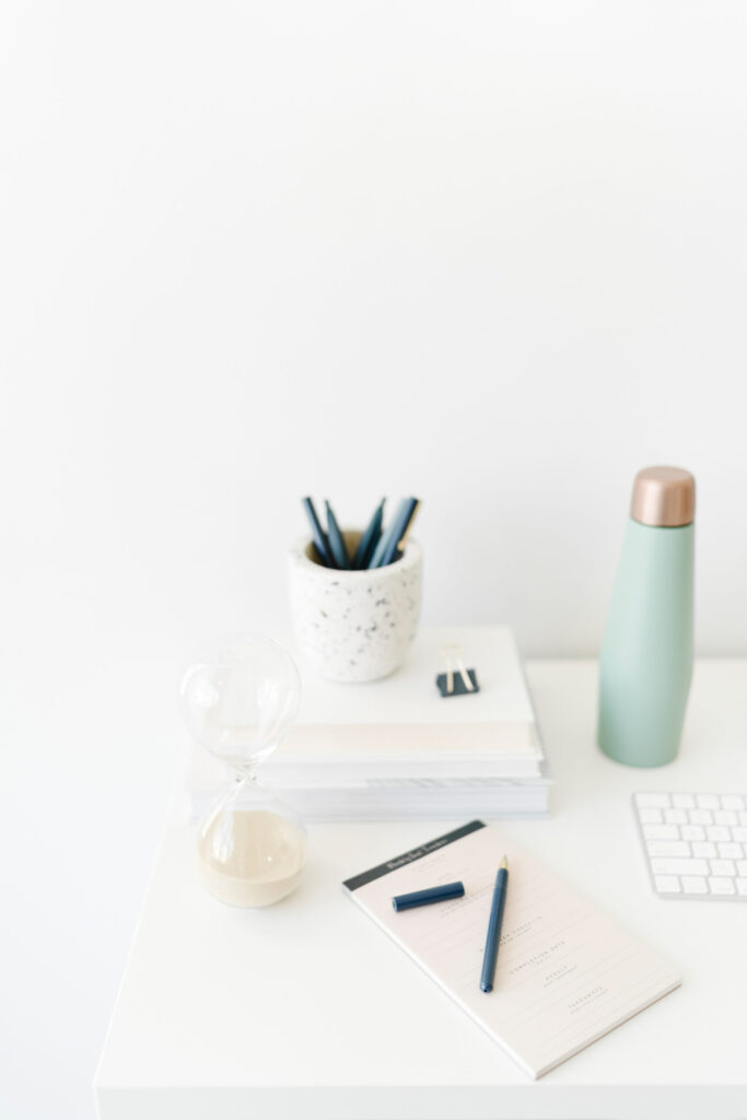 A white desk with an hourglass, planning worksheet, blue pens, stacked notebooks, keyboard, and mint-green water bottle.