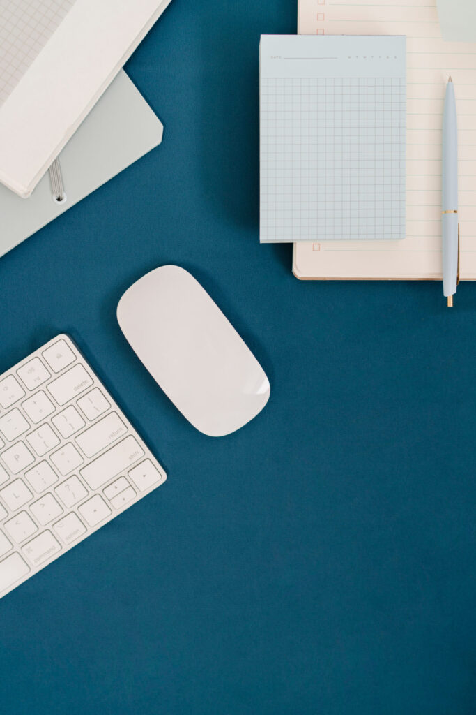 A white keyboard and mouse beside blue planning pads, notebooks, and a pen arranged on a deep blue desk.