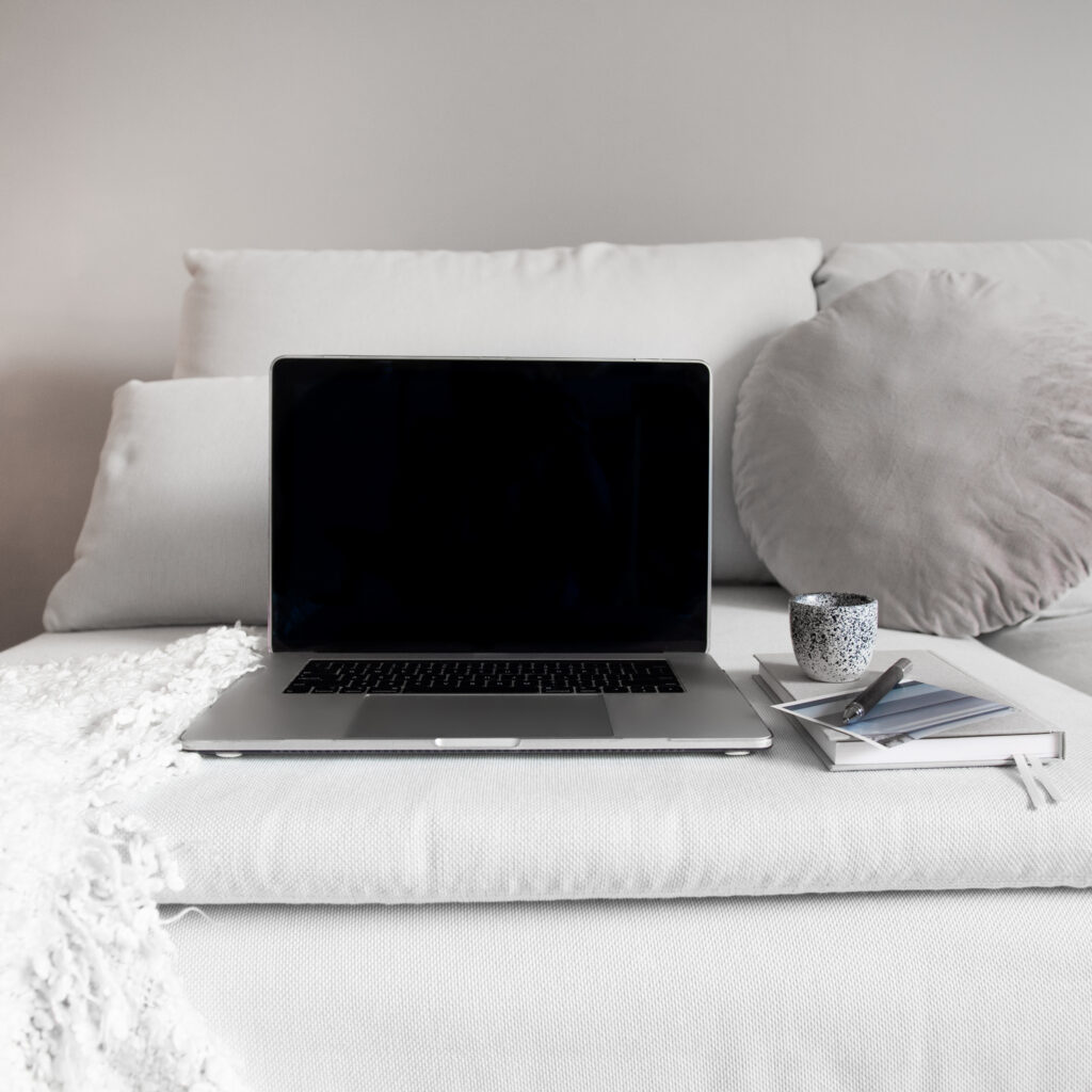 An open laptop sits on a neutral sofa beside a notebook, color swatches, pen, and ceramic cup.