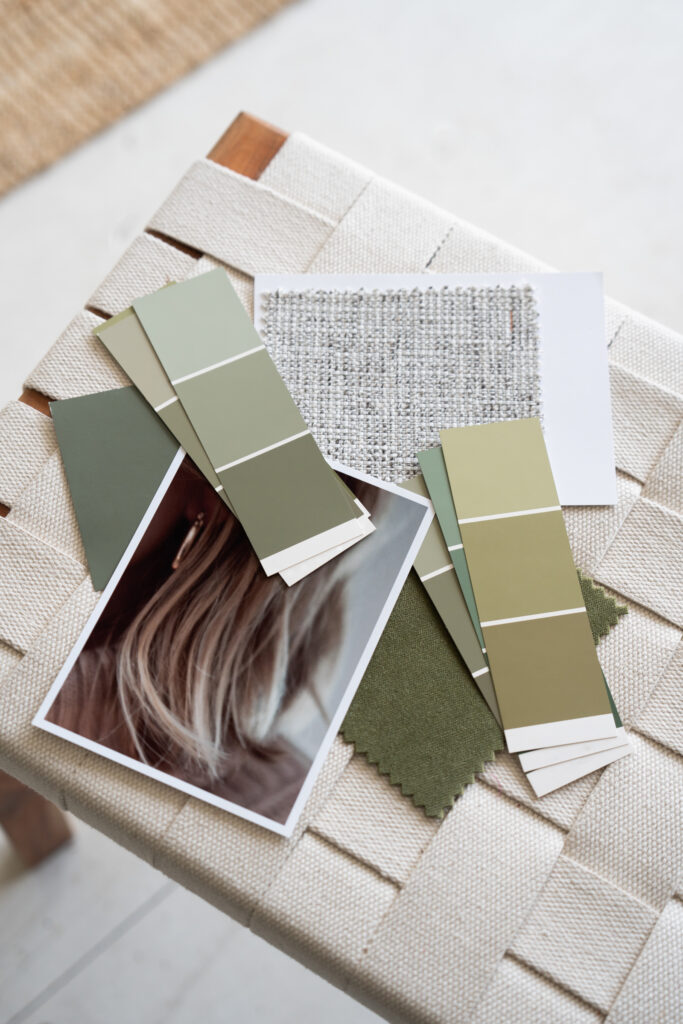 Green and neutral paint swatches, woven fabric samples, and a printed photograph arranged on a cream upholstered stool.