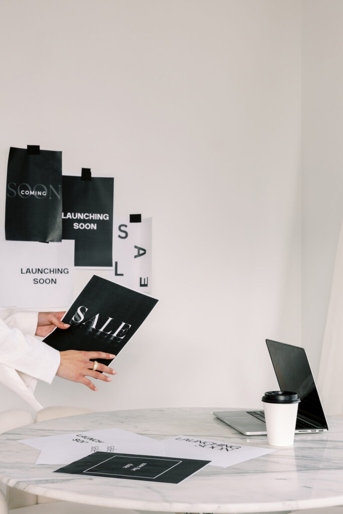Business owner holding a printed sale announcement beside a laptop, coffee cup, and launch materials.