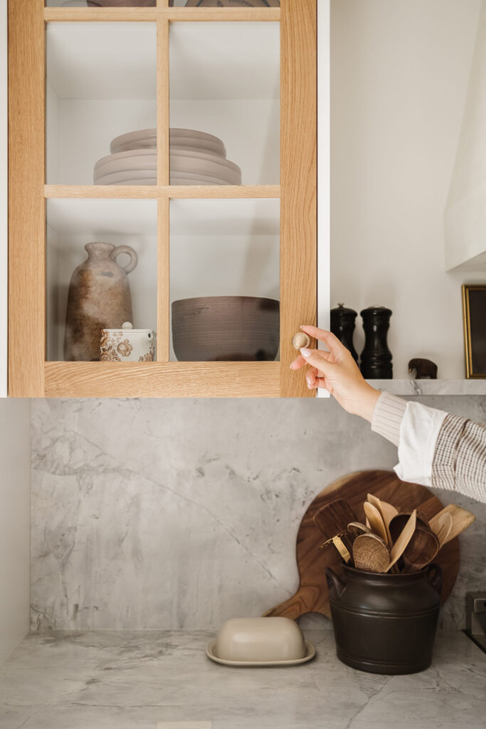 A person opening a glass-front kitchen cabinet filled with neutral dishes and serving pieces above a marble counter.