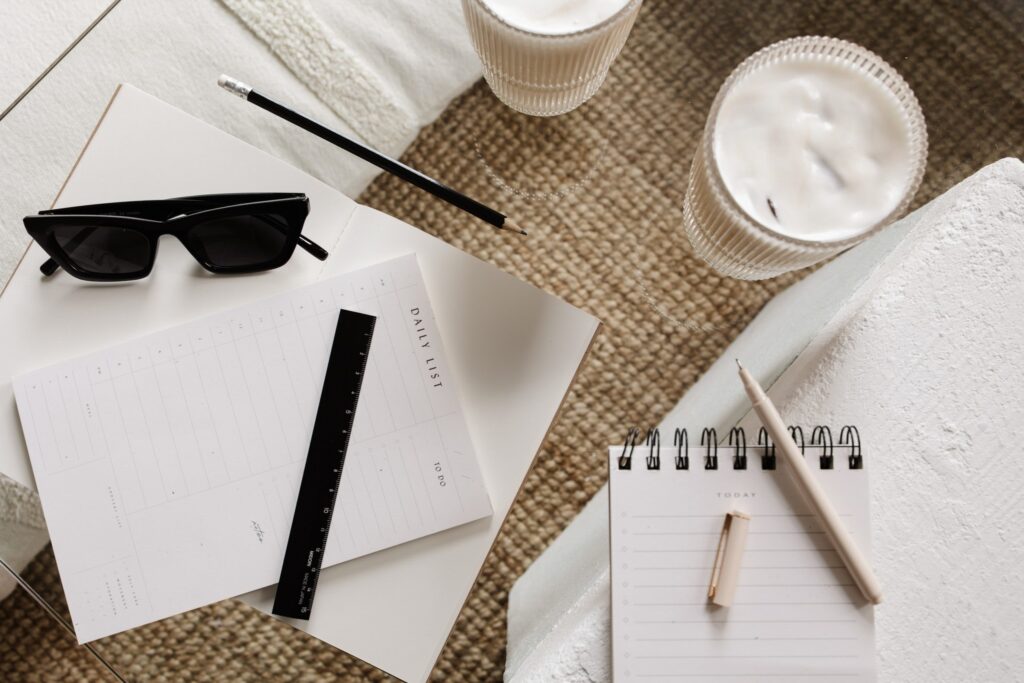 Daily planning sheets, notebooks, pens, black sunglasses, and two iced drinks arranged on a glass table.