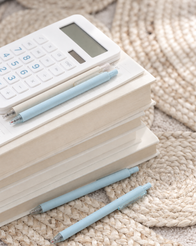 A white calculator and pale blue pens resting on a stack of cream books over a woven rug.