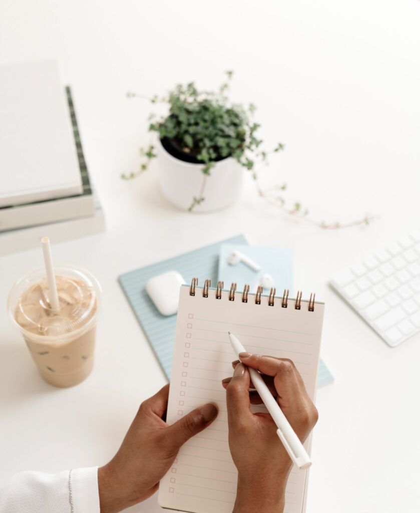Hands writing on a spiral checklist beside blue notebooks, a keyboard, earbuds, a plant, and iced coffee.