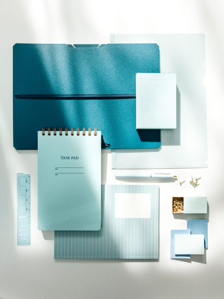 A spiral checklist, graph-paper card, sticky note, and pale blue pen arranged on a dark teal surface.