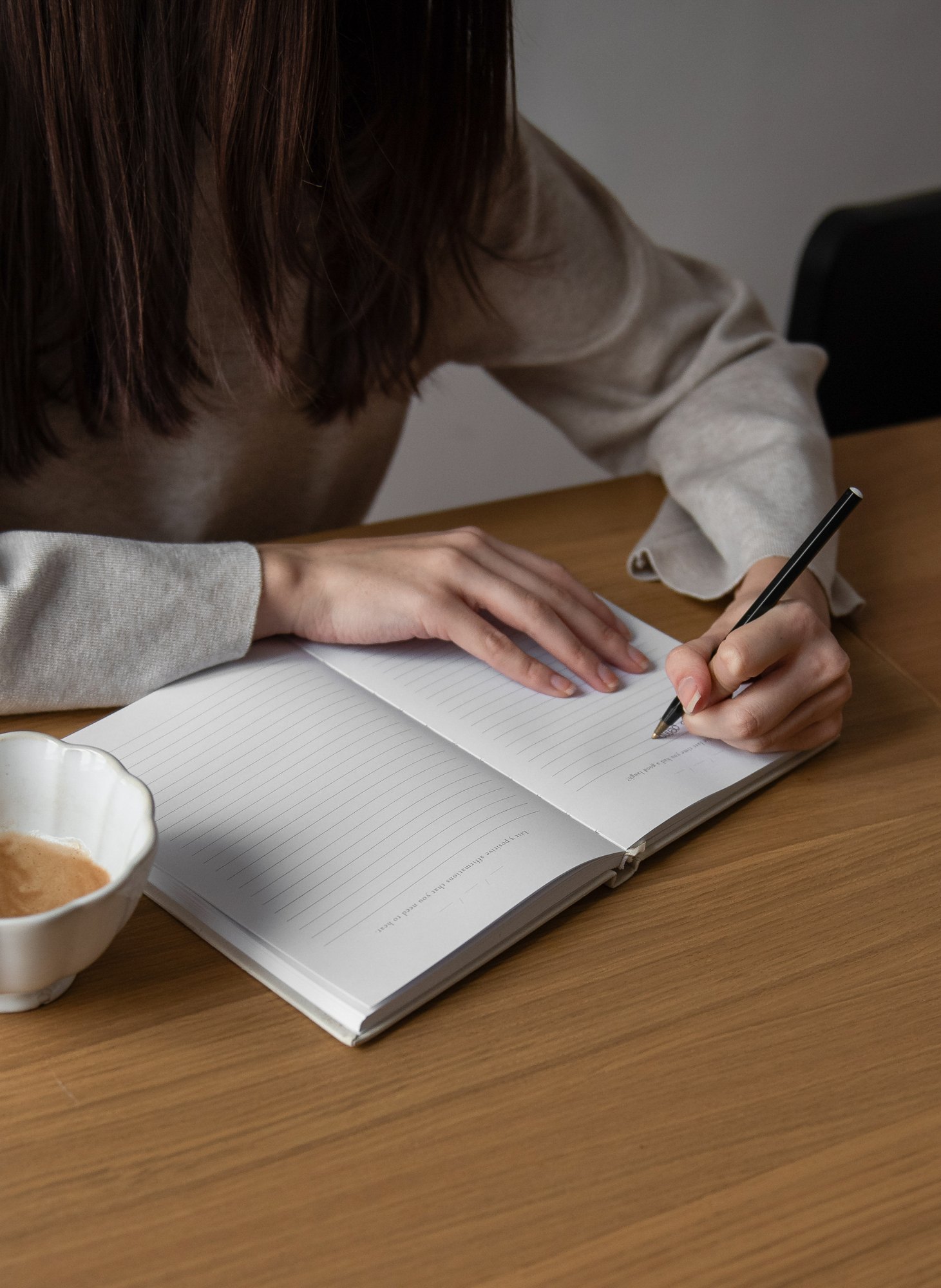 Person writing in a lined notebook beside a cup of coffee on a wooden table.